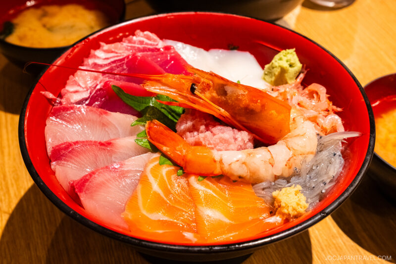 A colorful bowl of assorted sashimi, featuring shrimp, salmon, tuna, and other raw fish slices served on rice and garnished with wasabi and ginger—a true Numazu specialty. Miso soup is in the background.