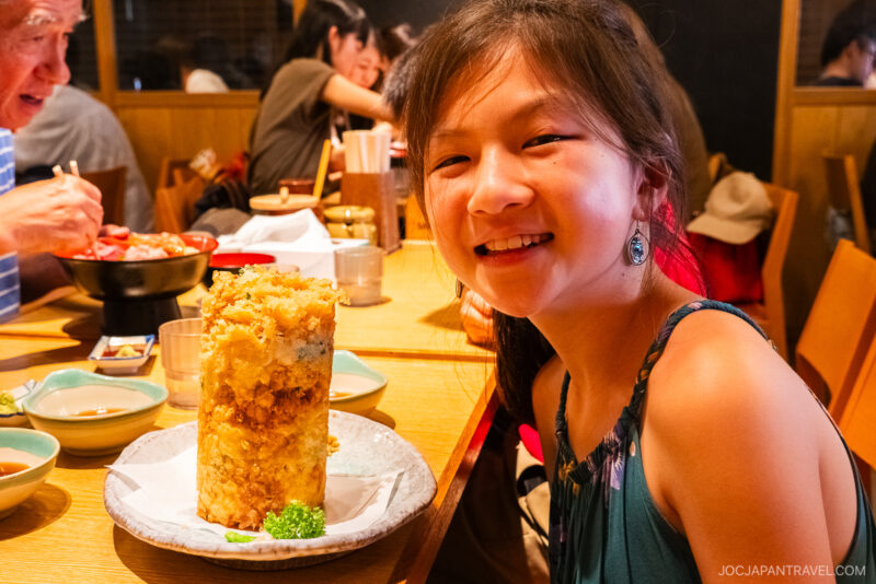 A smiling girl sits at a restaurant table in Numazu, with a tall, cylindrical tower of tempura in front of her, surrounded by small dishes and other diners in the background.