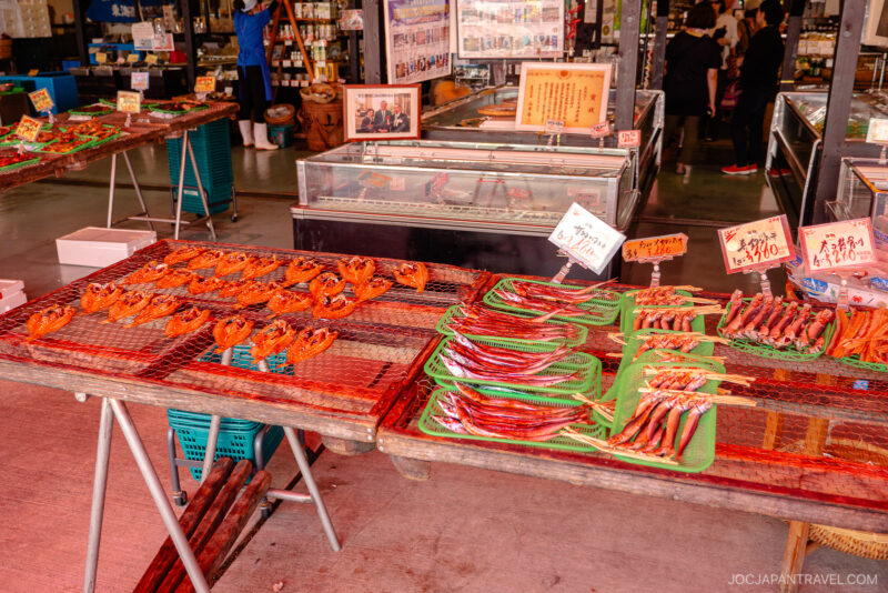 A market stall in Numazu displaying dried seafood, including reddish whole fish and long, thin fish arranged neatly on trays, with Japanese signs and people in the background.