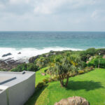 Seaside Hotel Yakushima – Ocean Views and Easy Access Near Miyanoura Port View of a grassy lawn with palm trees at Seaside Hotel Yakushima, bordered by modern buildings, overlooking rocky shorelines and the ocean under a cloudy sky. Waves crash against the rocks in the distance.