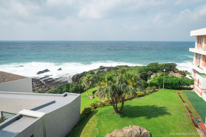 View of a grassy lawn with palm trees at Seaside Hotel Yakushima, bordered by modern buildings, overlooking rocky shorelines and the ocean under a cloudy sky. Waves crash against the rocks in the distance.