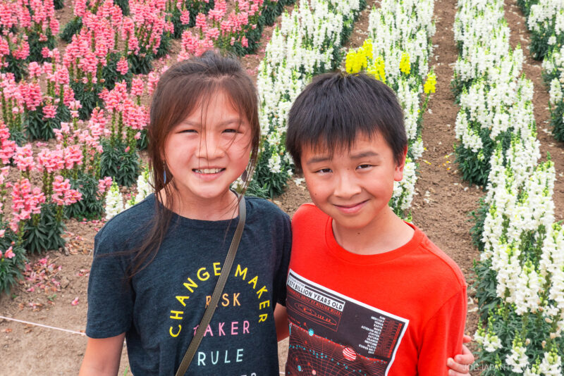 Two smiling children stand close together in front of rows of colorful blooming flowers at Shikisai no Oka. The girl wears a dark t-shirt with vibrant text, and the boy wears a red shirt, creating a cheerful and lively outdoor scene.