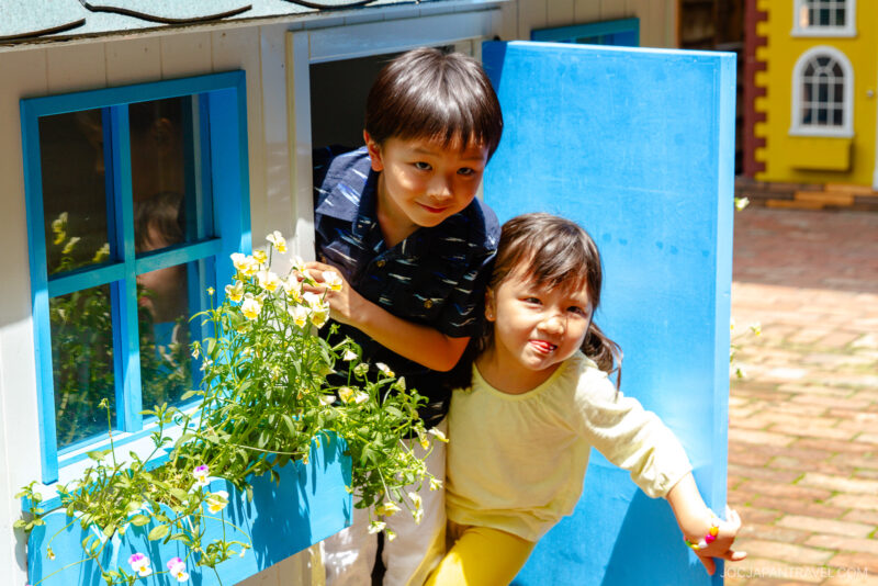 Two young children, a boy and a girl, lean out of a blue playhouse door at Shiroi Koibito Park. The boy smiles while the girl sticks out her tongue playfully. Flowers bloom in a window box beside them, with a brick path outside.