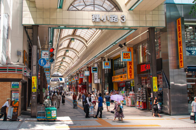 Covered shopping street in Japan with people walking, shops on both sides—including multiple ABC-MART stores—and a sign above reading 狸小路3. Tanukikoji Shopping Street is bright, lively, and bustling with shoppers.