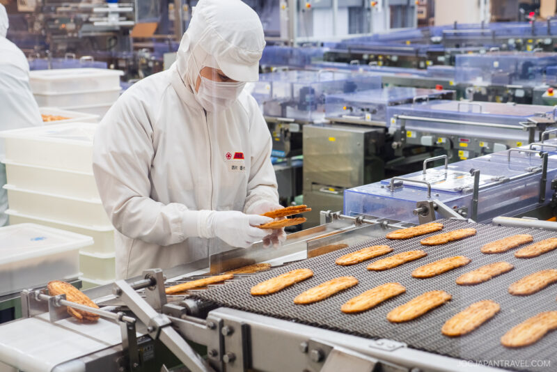 A worker in a white protective suit, mask, and gloves inspects pastries on a conveyor belt at the Unagi Pie Factory, surrounded by automated machinery in a modern food processing environment.