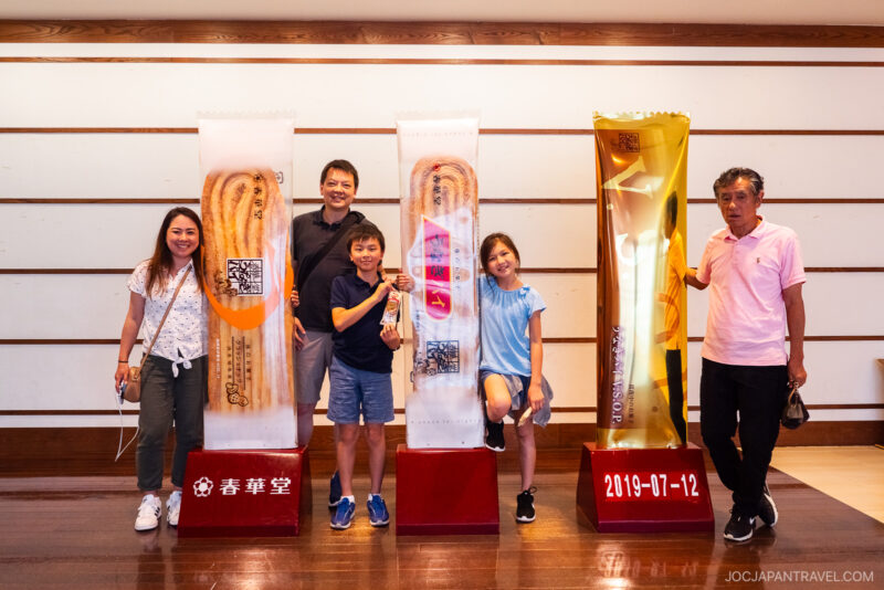 Five people stand smiling indoors by three large, colorful displays shaped like cookie sticks on red bases at the Unagi Pie Factory. The setting features a wooden floor and a white, striped wall background.