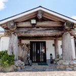 Yakudon Noodle House (屋久どん) – A Soul-Warming Lunch Stop in Yakushima A traditional Japanese building, Yakudon Noodle House features a rustic wooden entrance supported by large logs, surrounded by stone and greenery, set under a partly cloudy sky.