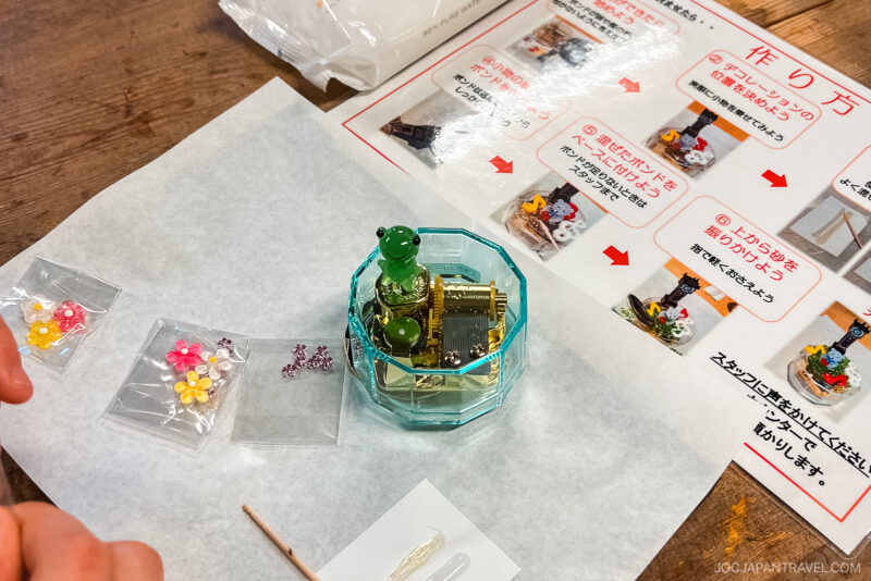 A crafting workspace at Yunokuni no Mori features a small frog figurine in a glass container, colorful flower beads in plastic bags, a toothpick, glue, and illustrated step-by-step instructions in Japanese on a wooden table.