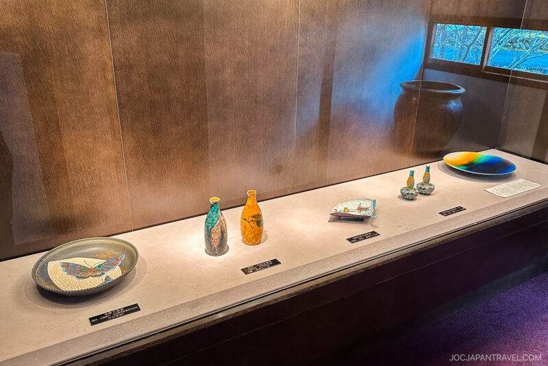 A display case with Japanese pottery items from Yunokuni no Mori, including plates, vases, and a bowl adorned with colorful designs, set on a white surface with labels and illuminated by soft lighting.