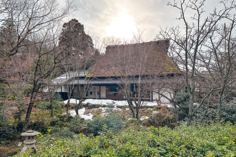 A traditional Japanese thatched-roof house in Yunokuni no Mori is surrounded by trees and greenery, with patches of snow on the ground and the sun shining through a cloudy sky in the background.
