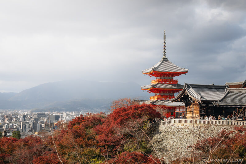 Red and orange kyoto autumn foliage surrounds the historic Kiyomizu-dera Temple, with the city and distant mountains visible under a cloudy sky in the background.
