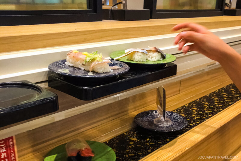 A hand reaches for a plate of sushi from a conveyor belt sushi restaurant. Several plates with sushi pieces glide by on the moving black track, offering a tempting selection.