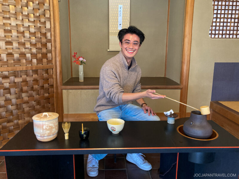 A smiling person wearing glasses sits at a traditional Japanese tea ceremony table in Uji, holding a bamboo ladle over a teapot, with tea utensils and cups arranged neatly in front of them.