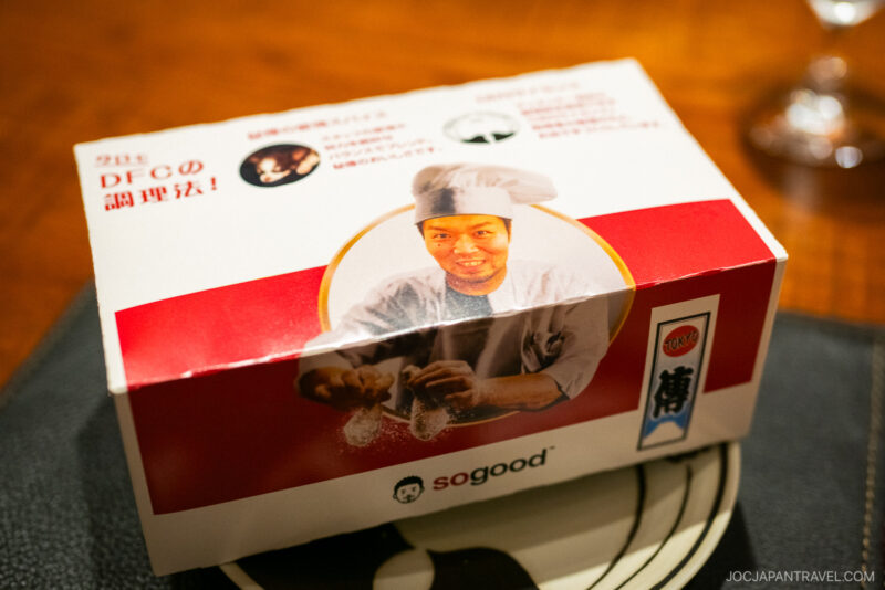 A red and white food box from den restaurant features a smiling chef sprinkling seasoning, Japanese text, and the sogood logo. The box sits on a placemat with a black and white design.