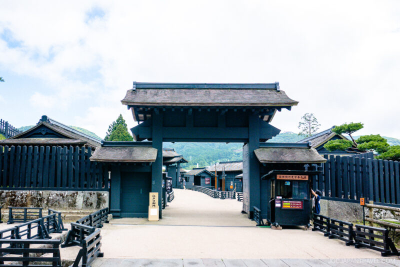 A traditional Japanese wooden gate and surrounding black wooden fences lead to an Edo-period samurai town recreation, inspired by the historic Hakone Checkpoint Museum, with mountains and trees in the background under a bright sky.