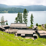 Hakone Checkpoint Museum (箱根関所) – A Glimpse Into Edo-Period Travel A traditional Japanese building complex with dark wooden roofs sits by a large lakeshore near the Hakone Checkpoint Museum, surrounded by lush greenery and tall trees, with forested hills rising in the distance.