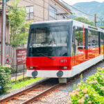 Hakone Freepass (箱根フリーパス) – The Best Way to Explore Hakone A red and white Hakone Tozan cable car travels up a steep track surrounded by greenery and buildings, with mountains visible in the background—a scenic ride accessible with the Hakone Freepass.
