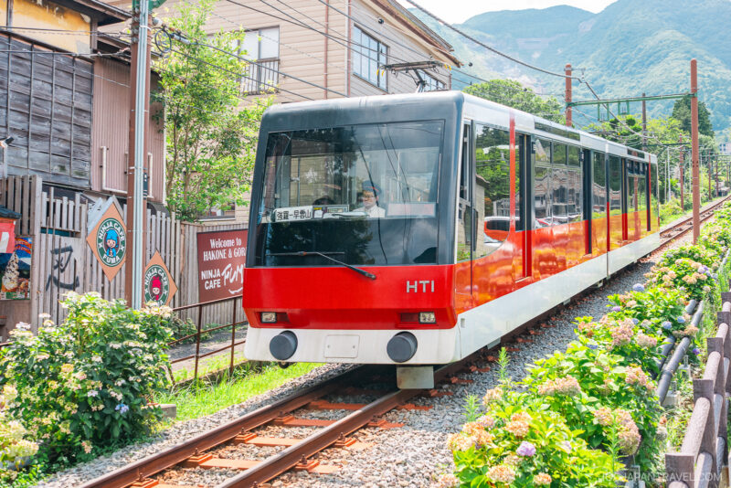 A red and white Hakone Tozan cable car travels up a steep track surrounded by greenery and buildings, with mountains visible in the background—a scenic ride accessible with the Hakone Freepass.