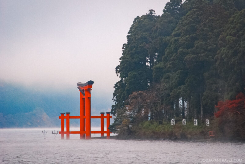 A bright red torii gate of Hakone Shrine stands in the water near the shore, surrounded by tall green trees and mist, creating a serene and atmospheric scene.