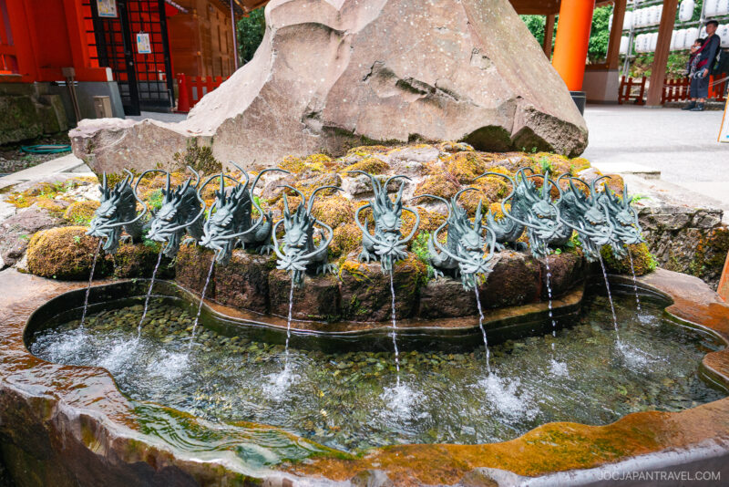 A row of dragon-shaped water spouts pours water into a stone basin at Hakone Shrine, surrounded by moss and vibrant orange pillars.