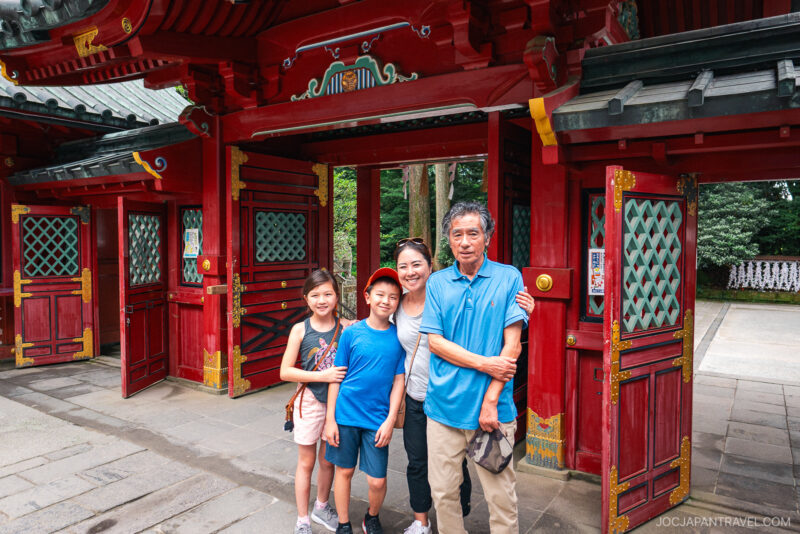 A smiling family of four poses in front of the bright red gate of Hakone Shrine, surrounded by lush greenery. Two casually dressed children stand in front, with two adults behind them, all enjoying their visit together.