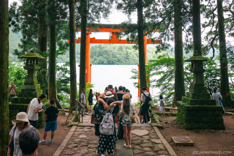 Tourists walk through a forested path toward the large red torii gate of Hakone Shrine by a lake, surrounded by tall trees and stone lanterns, with lush greenery in the background.