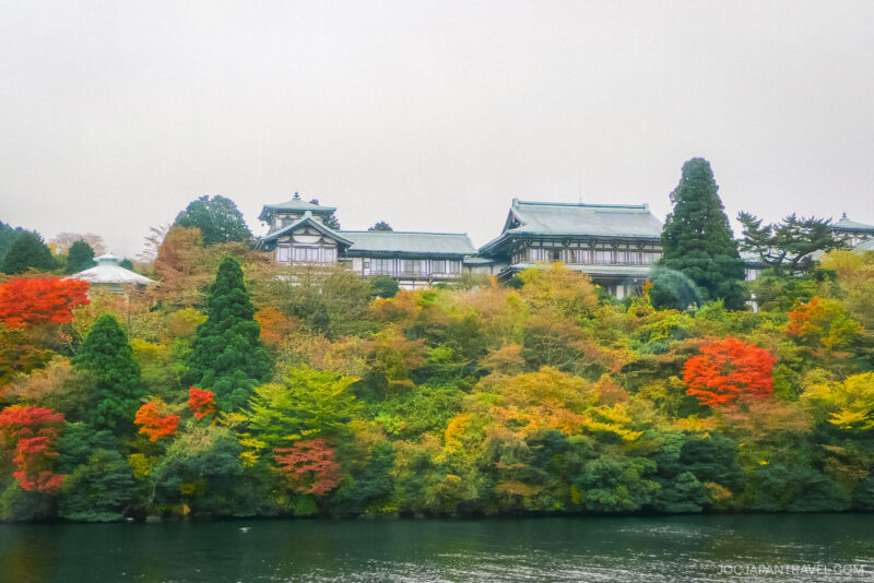 A traditional Japanese building sits atop a hill covered with colorful autumn trees in Hakone-en, overlooking a calm body of water under a cloudy sky.