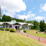 Hakone-en (箱根園) – Family Fun on the Shores of Lake Ashi A bright building with colorful cartoon banners sits among green lawns and trees under a blue sky at Hakone-en; people and horses are nearby, and outdoor umbrellas are set up on the grass.