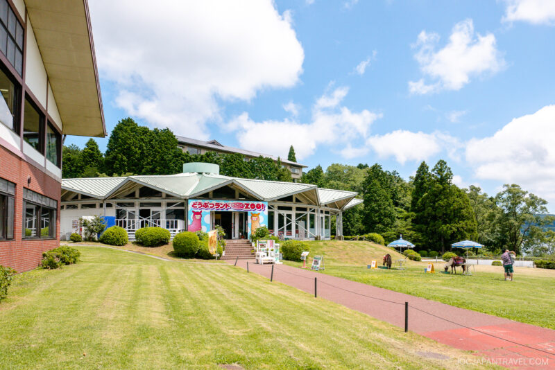 A bright building with colorful cartoon banners sits among green lawns and trees under a blue sky at Hakone-en; people and horses are nearby, and outdoor umbrellas are set up on the grass.