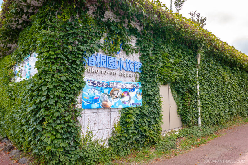 An ivy-covered building wall at Hakone-en features a sign reading Hakone-en Aquarium in English and Japanese, displaying images of aquatic animals and a welcome message. A door stands just beside the vibrant sign.