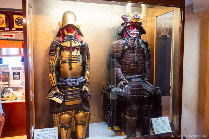 Two traditional Japanese samurai armors are displayed in a glass case at the museum. The armor on the right features a helmet with a gold crest, while both have intricate metal and leather detailing, reminiscent of those found at Hamamatsu Castle.