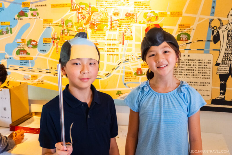 Two children wearing traditional Japanese helmets and holding a sword smile at the camera, standing in front of a colorful illustrated map with Japanese text, likely near Hamamatsu Castle.