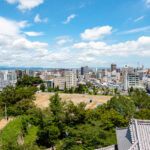 Hamamatsu (浜松市) – Music, Castles, and Coastal Adventures A cityscape view with modern buildings under a blue sky, scattered clouds, and a mix of green trees and open land in the foreground, with Hamamatsu Castle rising gracefully amidst the urban landscape.