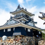 Hamamatsu Castle (浜松城) – Home of Tokugawa Ieyasu and the Castle of Success A traditional Japanese castle, like Hamamatsu Castle, with white walls and black tiled roofs, stands atop a stone foundation beneath a blue sky with light clouds. Some trees and a few people are visible near the entrance.