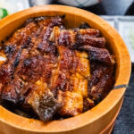 Hamamatsu Unagi – Japan’s Freshwater Eel Capital A close-up of a wooden bowl filled with Hamamatsu unagi—grilled eel glazed in sauce over rice—accompanied by cucumber slices, sliced green onions, and soup in the background.