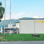 Hikari Miso Factory Tour A white industrial building with HIKARI MISO written in yellow stands on green grass, with flagpoles in front and mountains visible in the background—a perfect spot to begin your hikari miso factory tour under a blue sky.