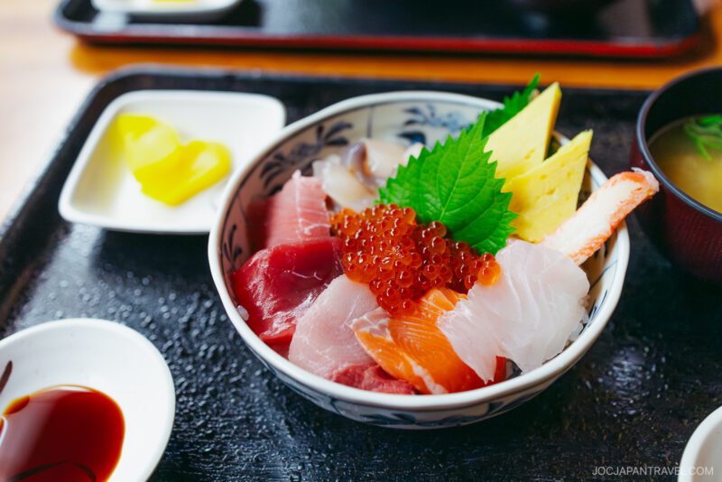 A bowl of Japanese chirashi sushi from Yoichi with assorted sashimi, fish roe, tamago, shiso leaf, and crab on rice, served alongside pickles, dipping sauce, and miso soup on a sleek black tray.
