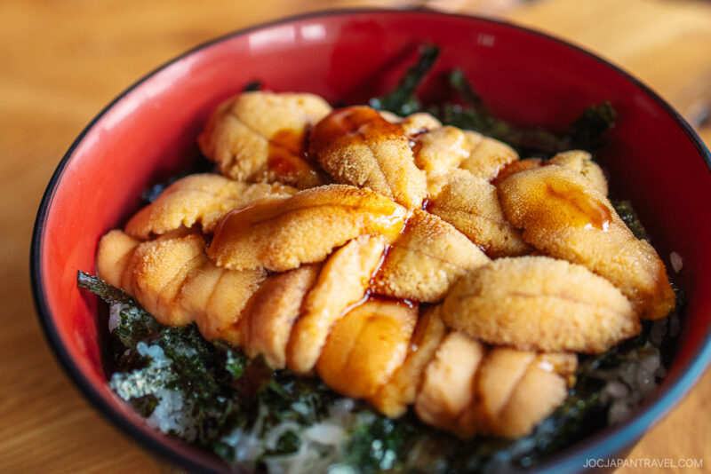 A close-up of a bowl of uni donburi from Yoichi, featuring fresh sea urchin over white rice with seaweed, drizzled with soy sauce, served in a red and black bowl.