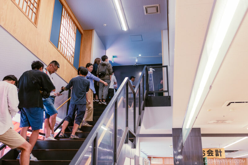 A group of people stand in line on a staircase inside a modern, yoichi-inspired building with blue and wooden walls, waiting to reach the upper floor.