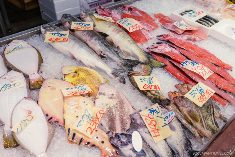 Fresh fish and seafood displayed on ice at a yoichi market stall, with handwritten price tags in Japanese attached to each type of fish. The selection includes various whole fish and fillets.