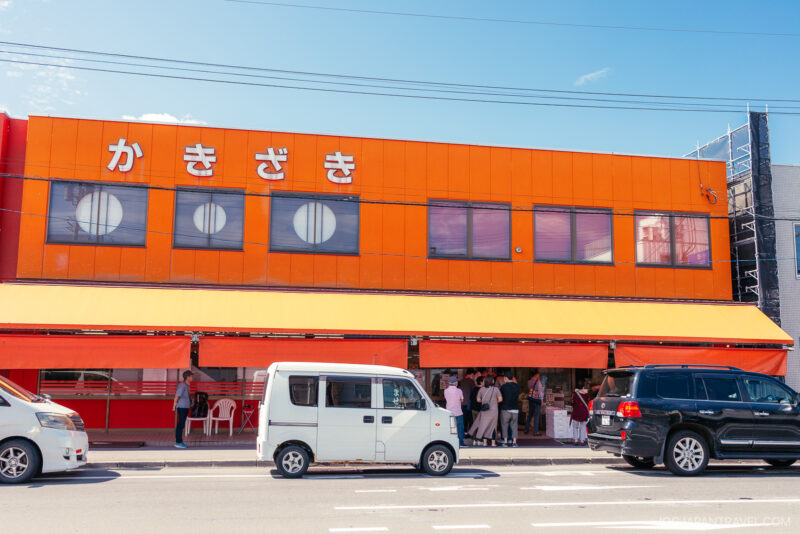 An orange two-story yoichi building with large Japanese characters on the facade. Several cars are parked outside, and people gather under the orange awning on a sunny day.