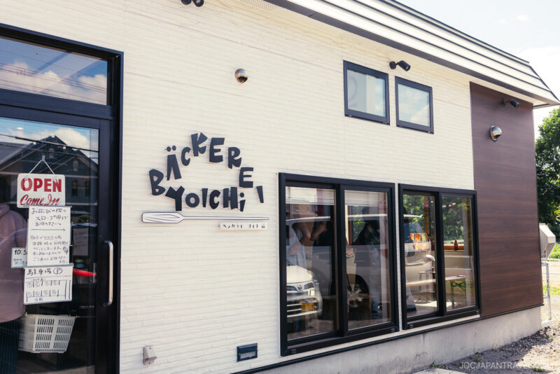 A bakery named Bäckerei Yoichi features a white exterior wall and a sign with "Yoichi." The shop window reflects trees and someone in a hat, while an OPEN sign and business hours are displayed near the entrance.
