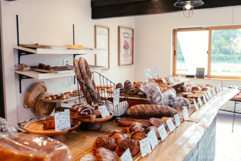 A bakery counter displays various freshly baked breads and pastries, with handwritten signs—one featuring Yoichi’s special loaf. Shelves in the background hold more bread, as sunlight streams in through a window.