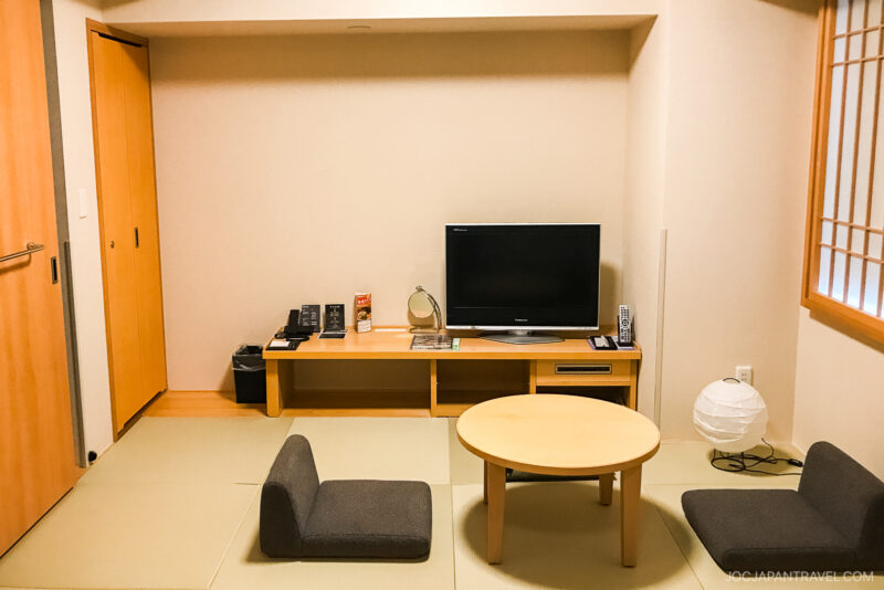 A minimalist Japanese-style room at Hotel Dormy Inn Kumamoto features tatami mats, a round wooden table, two floor chairs, a TV on a low stand, and a paper lantern by the shoji screen window.
