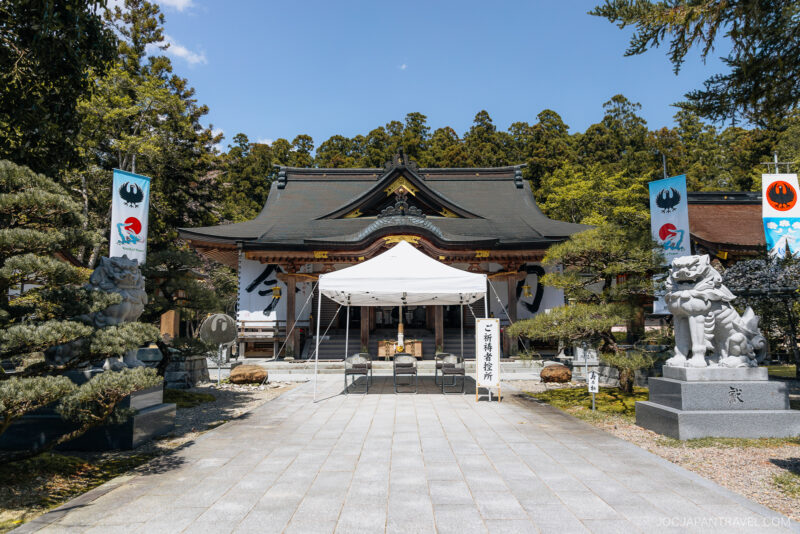 A traditional Japanese shrine, Kumano Hongu Taisha, with a dark tiled roof, stone statues, and a white tent in front. Tall banners with symbols stand on each side, surrounded by green trees under a clear blue sky.