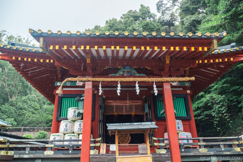 A traditional Japanese shrine, Kunozan Toshogu Shrine features a red wooden structure, ornate roof, green shutters, and decorative barrels, all surrounded by lush greenery.