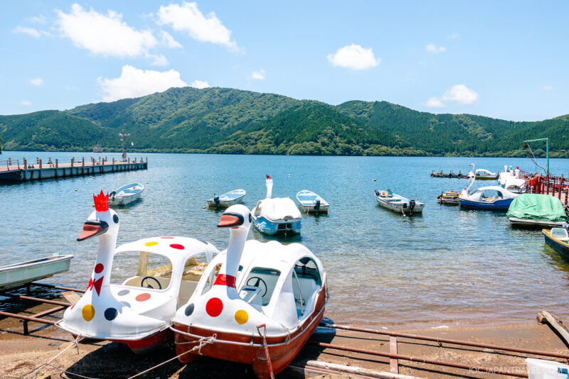 Colorful swan-shaped pedal boats sit on the shore of Lake Ashi, with several small boats floating nearby. A dock extends into the clear blue water, and green mountains rise in the background under a sunny sky.