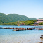 Lake Ashi (芦ノ湖) Travel Guide: Pirate Ships, Shrines, and Scenic Views A clear Lake Ashi with wooden docks, surrounded by green hills. A large, colorful ship resembling a pirate ship is docked near buildings in the background under a bright blue sky.