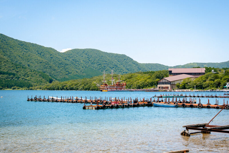 A clear Lake Ashi with wooden docks, surrounded by green hills. A large, colorful ship resembling a pirate ship is docked near buildings in the background under a bright blue sky.