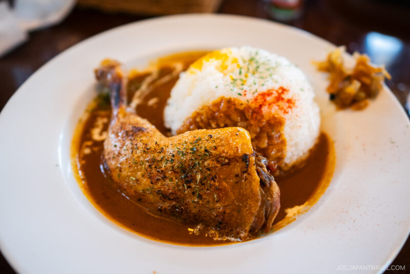A plate of Japanese curry rice topped with a seasoned chicken leg, served with a mound of white rice and herbs, evokes flavors as comforting as a meal enjoyed by scenic Lake Ashi, accompanied by pickled vegetables on the side.